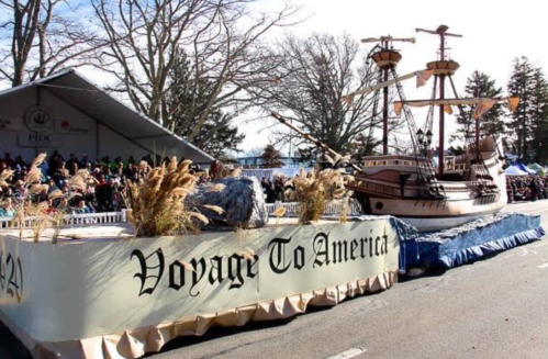 A parade float featuring a ship with "Voyage To America" displayed, surrounded by spectators and trees.