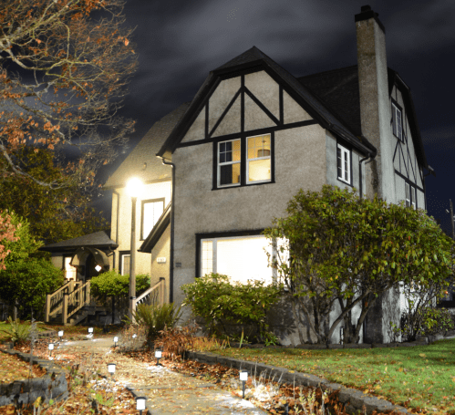 A charming two-story house at night, illuminated by warm lights, surrounded by trees and a well-lit pathway.
