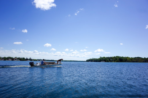 A boat glides across a calm blue lake under a clear sky with a few clouds and green trees in the background.