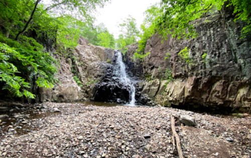 A serene waterfall cascading down rocky cliffs, surrounded by lush green trees and a stony riverbed.