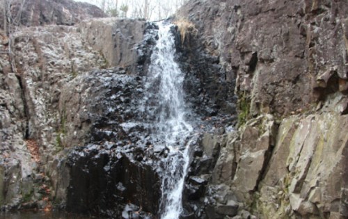 A small waterfall cascading over dark, rocky terrain surrounded by sparse trees and autumn foliage.