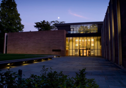 Modern building exterior at dusk, featuring large glass windows and a moonlit sky. Greenery in the foreground.