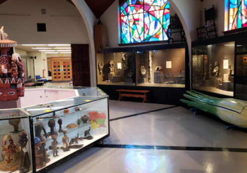 A museum interior featuring display cases with artifacts, colorful stained glass windows, and various sculptures.