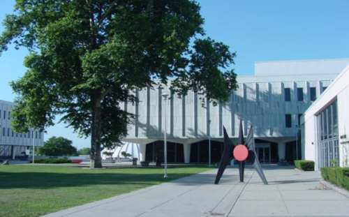 A modern building with a large sculpture in front, surrounded by green grass and trees under a clear blue sky.