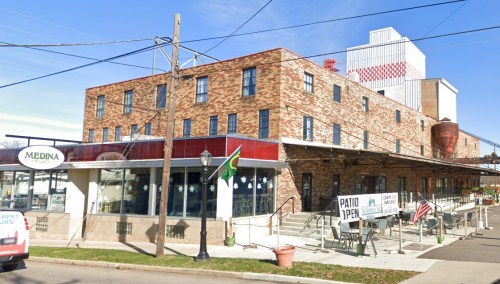 A brick building with large windows, featuring a patio area and flags, set against a clear blue sky.