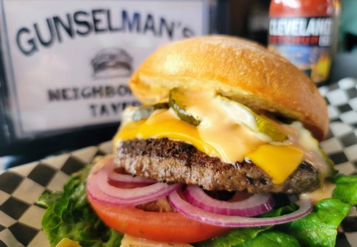 A close-up of a juicy burger with cheese, pickles, and onions, served on a bun with lettuce, next to a drink.