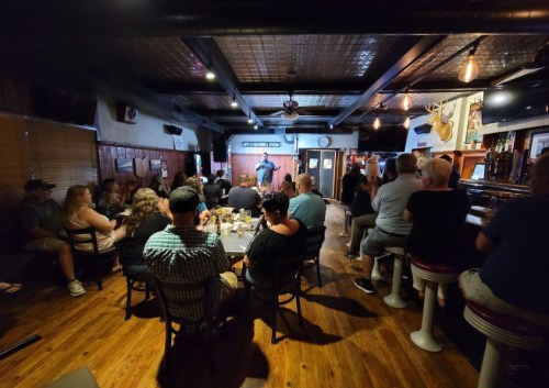 A comedian performs on stage in a lively bar, with an audience seated at tables and the bar counter.
