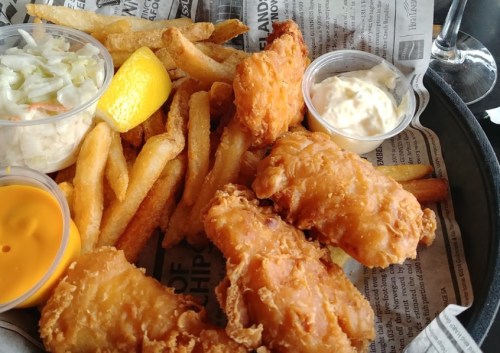 A plate of crispy fried fish, French fries, coleslaw, lemon, and dipping sauces on a bed of newspaper.
