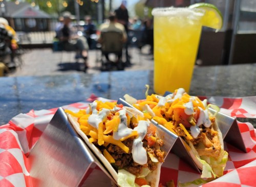 Two tacos with meat, cheese, and toppings in a red-checkered basket, with a yellow drink and outdoor seating in the background.
