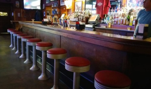 A bar with a polished wooden counter and red cushioned stools, lined up against a wall of bottles and mirrors.