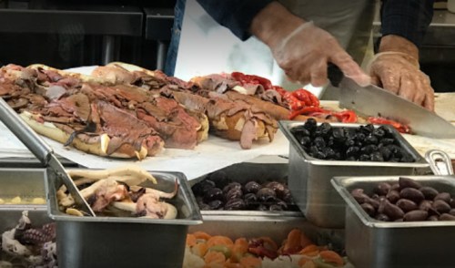 A chef slices meat on a countertop, surrounded by various ingredients like olives, mushrooms, and roasted peppers.