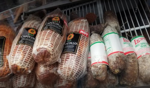 Various types of packaged cured meats displayed in a refrigerated case, including capocollo and salami.