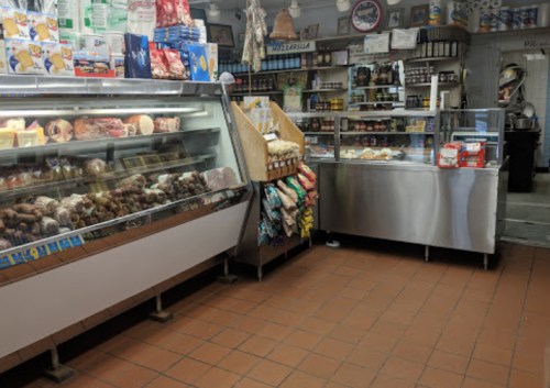 A cozy deli interior with display cases of meats and cheeses, shelves of canned goods, and a tiled floor.