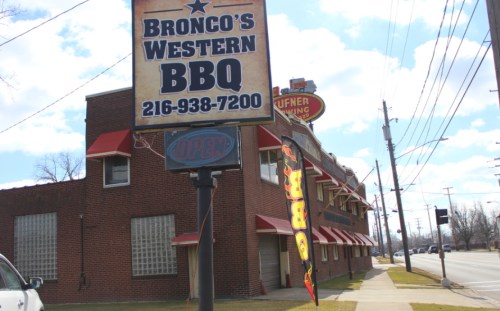Sign for Bronco's Western BBQ with "Open" banner outside a brick building on a sunny day.