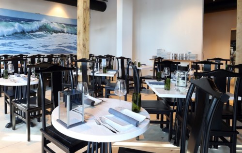 A modern restaurant interior with black chairs and white tables, featuring a coastal mural and neatly set dining areas.