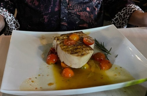 A plate featuring grilled fish topped with cherry tomatoes and garnished with rosemary in a flavorful sauce.