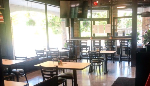 A bright, empty restaurant interior with tables and chairs, large windows, and a TV mounted on the wall.