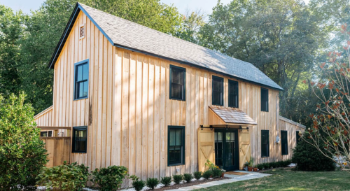 A modern wooden house with large windows, a sloped roof, and a welcoming entrance surrounded by greenery.