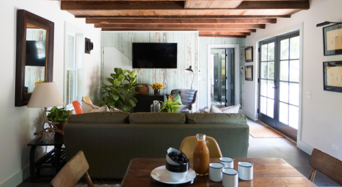 Cozy living room with wooden beams, a green sofa, plants, and a dining table set for breakfast. Natural light from windows.