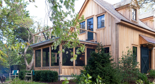 A wooden house with a screened porch, surrounded by greenery and trees, showcasing a cozy, rustic design.