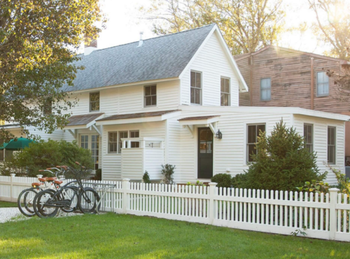 A charming white house with a picket fence, surrounded by greenery and bicycles parked in the front yard.