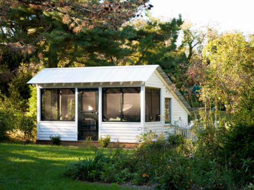 A small white cottage with a metal roof, surrounded by greenery and flowers, featuring large screened windows.