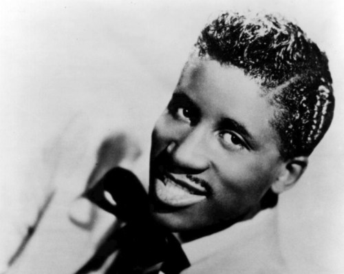 A smiling man with a stylish hairstyle, wearing a suit and bow tie, poses for a black-and-white photograph.