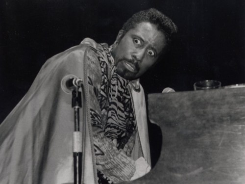 A musician in a dramatic pose at a piano, wearing a patterned outfit and a cape, with an intense expression.