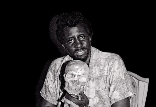 A man in a patterned shirt holds a skull, sitting against a dark background.