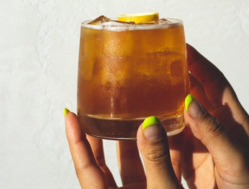 A close-up of a hand holding a glass of amber-colored cocktail with ice and a lemon slice.