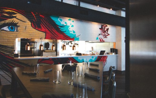 Modern restaurant interior featuring a vibrant mural, open kitchen, and stylish dining setup with glassware and napkins.