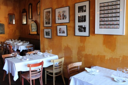 Cozy restaurant interior with wooden chairs, white tablecloths, and framed photos on a warm, textured wall.