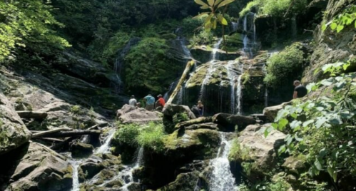 A scenic waterfall surrounded by lush greenery, with people exploring the rocky terrain nearby.