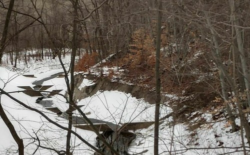 A winding creek flows through a snowy landscape, surrounded by bare trees and patches of brown leaves.