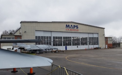 Exterior of MAPS Air Museum featuring a large hangar and military aircraft on display outside. Overcast sky.