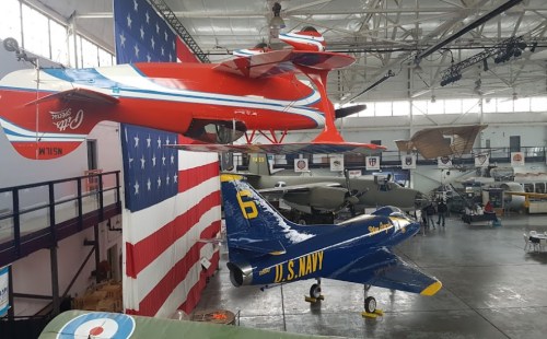 Two vintage aircraft hang in a museum, with a large American flag backdrop and various planes in the background.