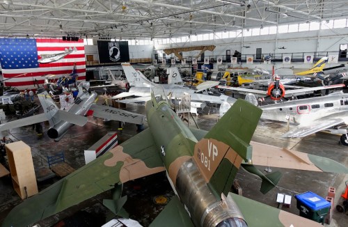 A spacious hangar filled with various aircraft, including military planes, under a large American flag backdrop.