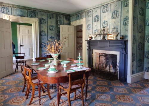 A vintage dining room with a wooden table set, blue wallpaper, and a fireplace, featuring decorative plates and a vase.