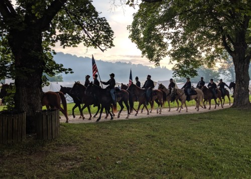 A group of riders on horseback, some carrying flags, passes through a tree-lined path in a scenic outdoor setting.