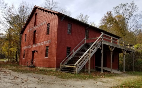 Red wooden building with a staircase, labeled "Carriage Manufactory," surrounded by trees in a rural setting.