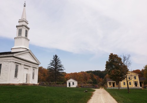 A white church stands beside a yellow house, surrounded by autumn trees and a gravel path.