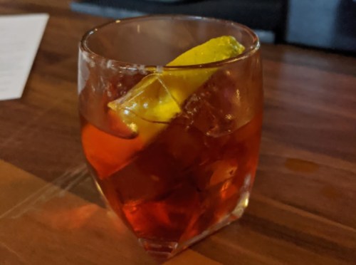 A glass of amber-colored cocktail with ice and a lemon wedge on a wooden surface.