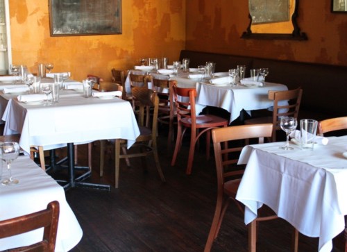 A cozy restaurant interior with wooden chairs and tables set with white tablecloths and glassware.