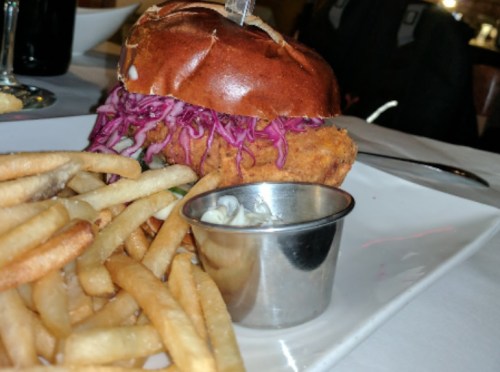 A crispy chicken sandwich with purple cabbage on a pretzel bun, served with a side of fries and dipping sauce.