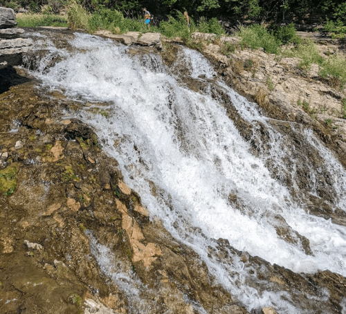 A cascading waterfall flows over rocky terrain, surrounded by lush greenery and sunlight.