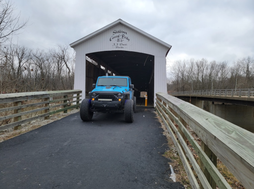 A blue Jeep parked at the entrance of a covered bridge, surrounded by bare trees and a cloudy sky.