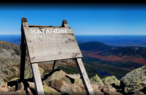 Sign marking Katahdin summit with a clear blue sky and mountainous landscape in the background.