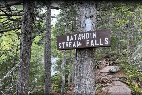 Sign for Katahdin Stream Falls surrounded by trees in a natural setting. Pathway visible leading into the woods.