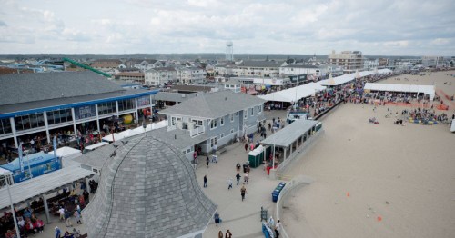 Aerial view of a beach boardwalk with crowds, shops, and a sandy beach under a cloudy sky.