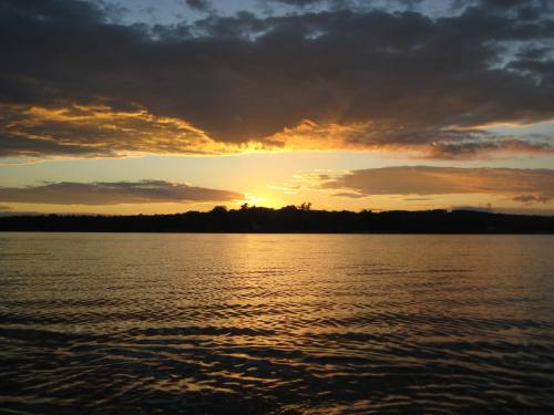 Sunset over a calm lake, with vibrant orange and yellow hues reflecting on the water and dark clouds in the sky.